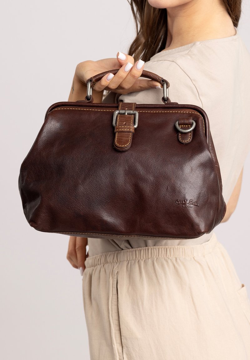 Femme tenant un sac à main en cuir brun foncé avec une boucle et des anneaux métalliques, portant un haut et un pantalon beiges devant un fond uni.