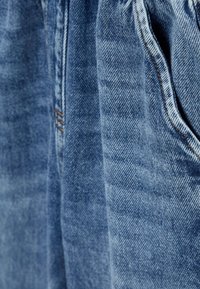Denimkangas vaaleansinisellä sävyllä, jossa on hieno haalistuminen. Siinä on ommeltuja saumoja ja taskuja. Kankaassa on hieman karkea ja kestävä rakenne.