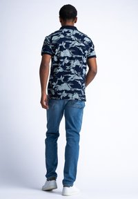 Námořnická modrá polokošile s krátkým rukávem, s jemným květinovým a horským vzorem, v kombinaci s světle modrými džínami a bílými teniskami.