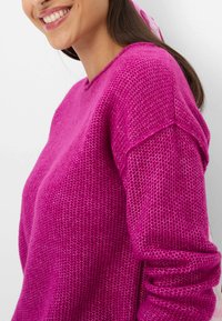 Fuchsia gestrickter Pullover mit strukturiertem Muster, V-Ausschnitt und überschnittenen Schultern, vor einem hellen Hintergrund präsentiert.
