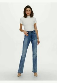 Jeans de mezclilla azules de corte acampanado con una cintura de altura media, estilo de cinco bolsillos, un ligero desgaste y un cierre clásico de botones. Combinados con una blusa blanca.