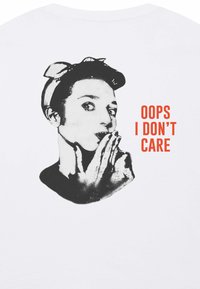 Balta marškinėliai su juodu piešiniu, vaizduojančiu asmenį su galvos juosta, burna uždengta ranka. Tekstas skaito "OOPS I DON'T CARE" oranžine spalva.