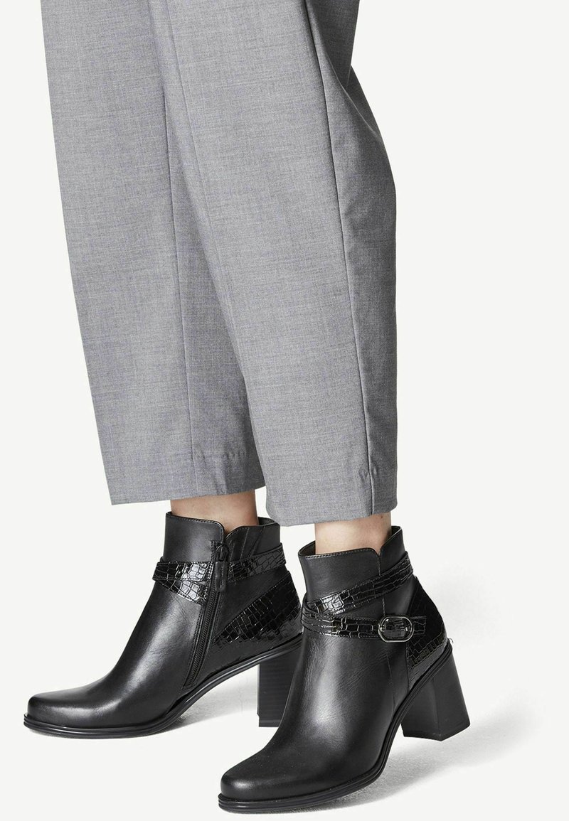 Bottes en cuir noir montant à la cheville avec talons blocs et détails de sangles texturées portées avec un pantalon gris coupé court.