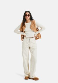 Beige Fleece-Weste über einem cremefarbenen strukturierter Oberteil, kombiniert mit hellbeigen, weit geschnittenen Hosen und braunen Slippern. Runde dunkle Sonnenbrille inklusive.