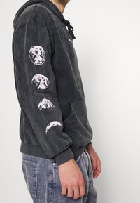 Felpa con cappuccio grigio scuro realizzata in tessuto morbido, con tasca frontale e grafiche rosa delle fasi lunari sulla manica sinistra.