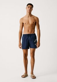 Pantaloncini da bagno blu navy con vita elastica e cordoncino, con testo stampato bianco sul lato. Abbinati a sandali beige chiaro.