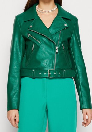 Woman wearing a green leather biker jacket with zipped pockets and belt, teal pants, and a double-strand pearl necklace.