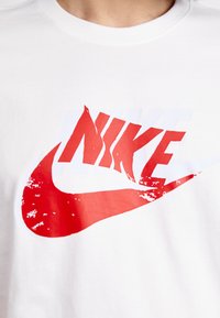 Baltas medvilninis marškinėliai su dideliu raudonu Nike logotipu su dėvėtumo akcentais, uždengtu baltu swoosh grafiku. Klasikinio apvalaus kaklo dizainas.