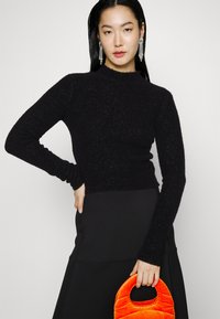 Schwarzer, strukturierten Langarmpullover mit einem Stehkragen, kombiniert mit einem schwarzen Rock und einer orangefarbenen, gesteppten Handtasche.