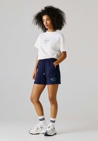 Bílé bavlněné tričko s potiskem loga, doplněné tmavě modrými šortkami s kapsami a nápisem "Red Bull Racing". Bílé ponožky s černými pruhy.