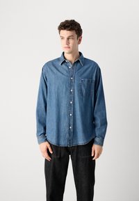 Camicia in denimazzurro medio con taschino e bottoni abbinati. Caratterizzata da maniche lunghe e orlo curvo. Abbinata a jeans scuri.