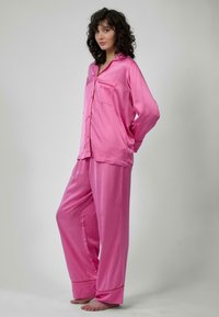 Set di pigiama in satin rosa composto da una camicia a maniche lunghe con bottoni, dotata di una tasca sul petto, e pantaloni larghi con gambe ampie e polsini.