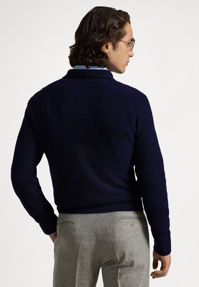 Marineblå strikket genser med ribbestruktur og krage, kombinert med lysegrå bukser med baklommer og metallbeslag.