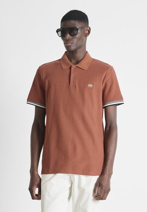 Homme portant un polo couleur rouille avec des bords de manches rayés, un pantalon blanc et des lunettes de soleil noires surdimensionnées sur fond blanc uni.