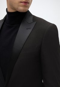 Zwarte smokingjas met satijnen revers, met daaronder een textuurrijke zwarte coltrui. Zakdetail zichtbaar aan de linkerkant.