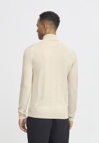 Pulóver de punto beige con cuello alto, mangas largas y una textura suave. Presenta un diseño ajustado y detalles de costura sutiles.