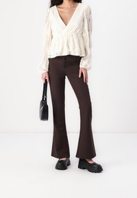 Blusa color crema con peplum, maniche lunghe trasparenti e dettagli in pizzo, abbinata a pantaloni svasati marroni scuri e scarpe con plateau nere.