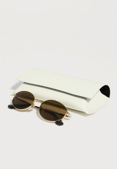 Οβάλ καφέ αποχρώσεων γυαλιά ηλίου με χρυσά πλαίσια που ακουμπούν δίπλα σε μια κλειστή εκρού δερμάτινη θήκη πάνω σε λευκή επιφάνεια.