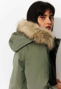 Parka verte avec une capuche doublée de fourrure, dotée d'une fermeture éclair et de boutons-pression. Le tissu a une texture lisse avec des coutures minimales.