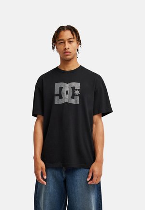 Jeune homme aux cheveux tressés, portant un t-shirt noir avec un grand logo DC blanc et un jean bleu, debout devant un fond clair uni.