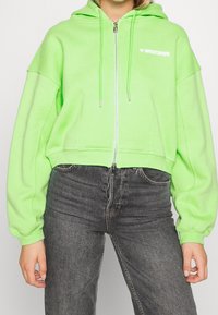 Felpa corta con cappuccio verde neon con coulisse, chiusura con zip e logo bianco sul petto a sinistra. Presenta una vestibilità ampia e maniche lunghe.