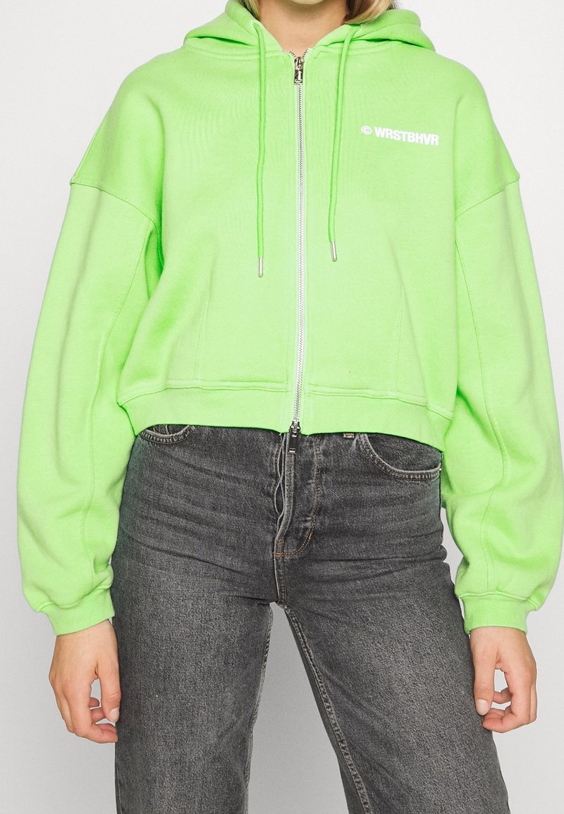 Felpa corta con cappuccio verde neon con coulisse, chiusura con zip e logo bianco sul petto a sinistra. Presenta una vestibilità ampia e maniche lunghe.