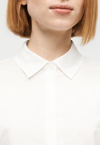 Λευκό πουκάμισο με κουμπιά και κλασικό γιακά, κατασκευασμένο από απαλό βαμβακερό ύφασμα με κλείσιμο με ένα κουμπί στον γιακά. Καθαρός και μίνιμαλ σχεδιασμός.