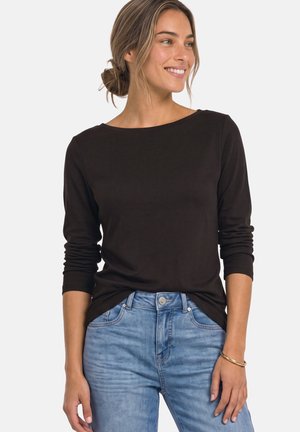 Femme aux cheveux tirés en arrière, portant un haut noir à manches longues et un jean bleu, souriante et regardant sur le côté sur un fond uni.