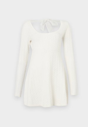 Hvid langærmet sweaterkjole med kabelstrikket struktur, åben ryg og sløjfedetalje ved halsen. A-linje silhuet.