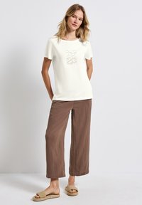 T-shirt blanc à manches courtes avec un design en relief ; associé à un pantalon large marron froissé et des sandales tissées à enfiler.