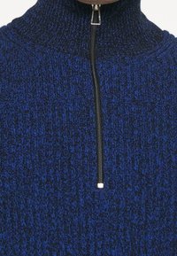 Strickpullover mit hohem Kragen, mit einem blau-schwarzen Muster und einem Reißverschluss vorne mit einem silbernen Ziehgriff. Weiche Textur.