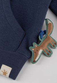 Marineblauer Pullover mit strukturiertem Rippenbündchen, versehen mit einem verspielten Stoff-Filtertag, der mit einem blauen Band befestigt ist. Das Etikett zeigt einen hellbraunen Farbklecks.