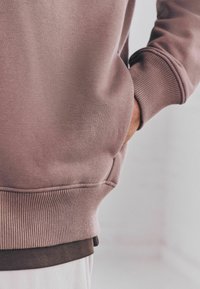Sudadera de color malva claro con puños y dobladillo acanalados, que presenta un bolsillo lateral y un aspecto superpuesto con una camiseta interior marrón oscuro. Textura suave.