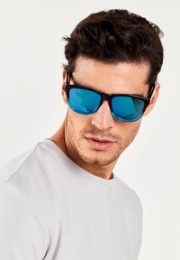 Ochelari de soare negri cu lentile reflective albastre, având un design elegant. Modelul poartă o cămașă scurtă, cu mânecă scurtă, texturată, de culoare gri deschis.