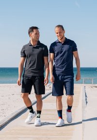 Due uomini camminano su un sentiero di legno vicino alla spiaggia. Uno indossa una polo grigio scuro e pantaloni corti neri; l'altro indossa una polo blu navy e pantaloni corti abbinati.