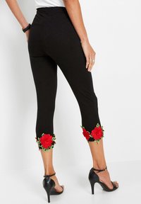 Donna che indossa leggings neri a vita alta con ricami di rose rosse vicino all'orlo, abbinati a sandali neri con tacco alto e cinturino alla caviglia.