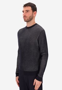 Uomo con capelli corti scuri e barba, che indossa un maglione a maniche lunghe a righe nere e grigie, in piedi con le mani rilassate ai lati.