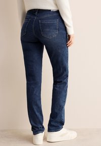 Jeans de mezclilla azul oscuro con ajuste slim, que presentan dos bolsillos traseros y detalles de costura sutiles, combinados con zapatillas blancas.