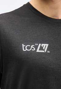 Κοντινό πλάνο ενός μαύρου υφασμάτινου πουκάμισου με τρύπες, με λευκή επιγραφή "tcs 20 L77 26" στο στήθος κοντά στο λαιμό.