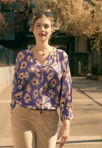 Blumenhemd mit Knopfleiste, aus lila Stoff mit weißen Gänseblümchen und roten Zentren. Helle beige Hosen mit Standardtaschen.
