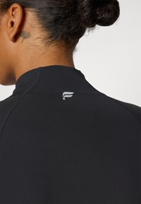 Crni sportski top s visokim ovratnikom, glatke teksture i malim logotipom na stražnjem dijelu vrata. Šavovi su vidljivi i uredno ušiveni.