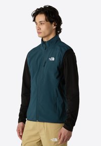 Teal softshell vest med høj krave, front lynlås og to lynlåsede lommer. Sorte lange ærmer kan ses nedenunder.