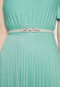 Robe plissée vert menthe avec ceinture assortie présentant une rangée centrale d’ornements de pierres précieuses scintillantes à la taille.