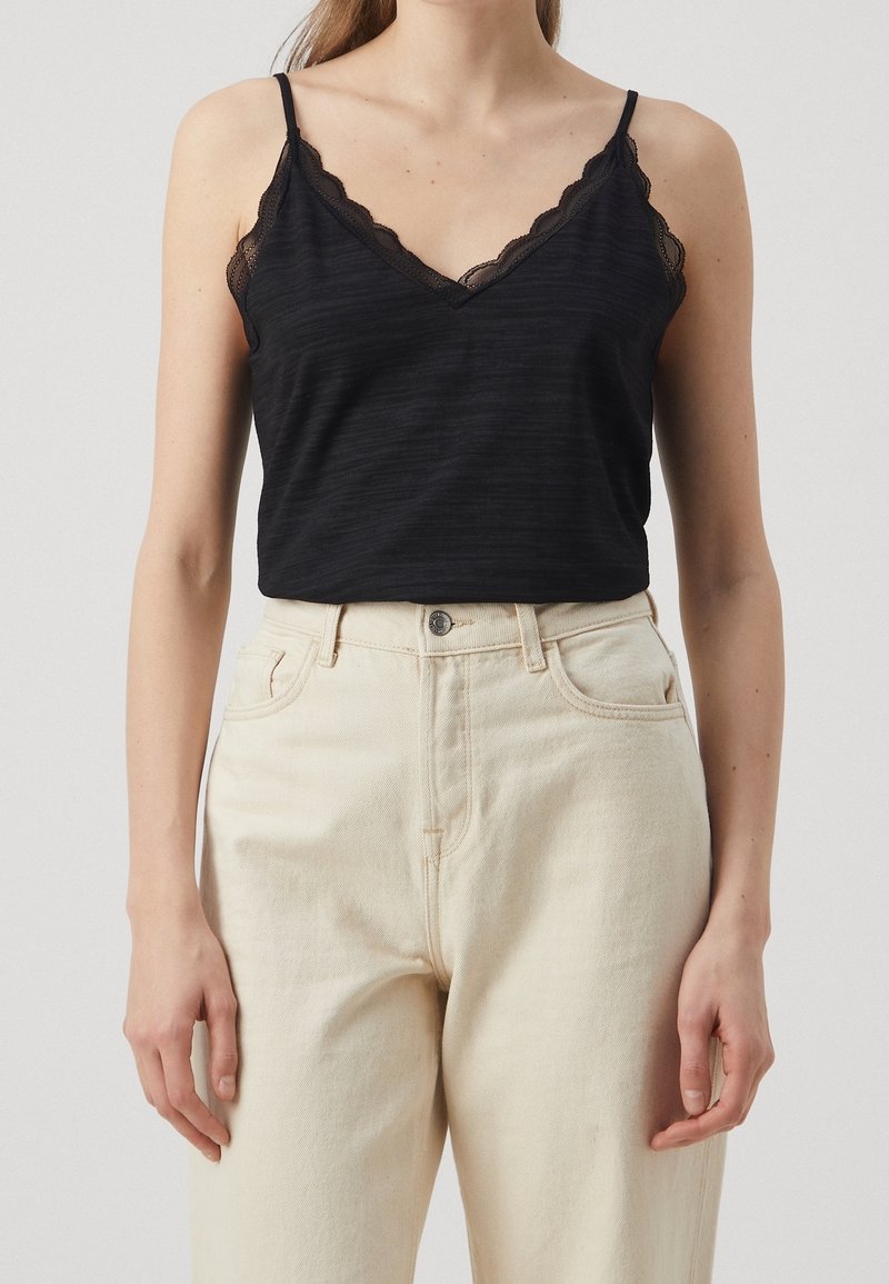 Černý top s výstřihem do V a krajkovým lemováním; vyroben z měkké, mírně texturované látky; nošen s krémovými kalhotami s vysokým pasem.
