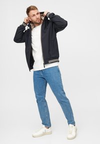 Veste bomber zippée bleu marine avec intérieur beige, poignets et taille côtelés ; portée avec un jean bleu clair et des baskets blanches.