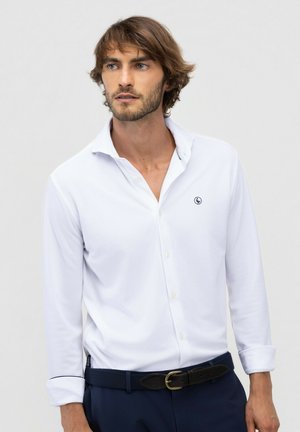 Hombre con pelo castaño, vestido con una camisa blanca abotonada y pantalones azul marino, mirando hacia un lado con un fondo liso y claro.