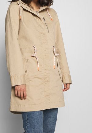 Femme portant un parka beige à capuche avec des cordons blancs et orange, fermeture éclair sur le devant, poches à rabat, et jean bleu, debout devant un fond gris clair.