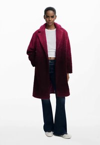 Bordo tekstūruotas paltas su įpjauta atvėrimo apykakle, viena sagomis užsegimu ir dviem šoninėmis kišenėmis, derinamas su platėjančiomis džinsinėmis ir baltais batais.
