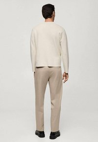 Homme portant un haut à manches longues crème, un pantalon beige et des chaussures noires, debout de dos sur un fond clair uni.