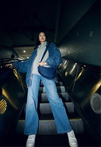 Giacca di jeans blu sopra un hoodie grigio, abbinata a jeans svasati blu chiaro e sneakers bianche, in piedi su una scala mobile in uno spazio poco illuminato.